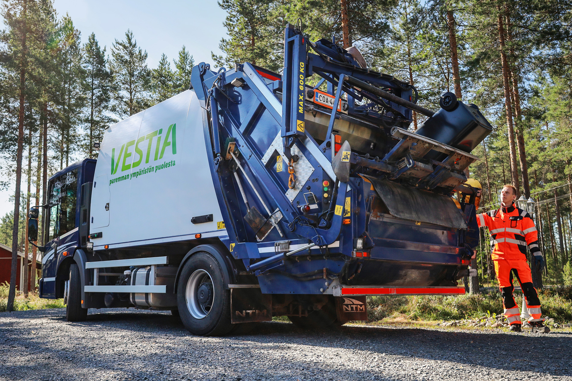 Jäteautonkuljettaja tyhjentämämässä jäteastiaa Vestian jäteautoon. Taustalla metsää ja punainen omakotitalo.