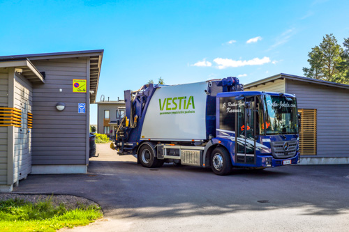 Vestian jäteauto taloyhtiön pihassa. Taustalla näkyy harmaita autokatoksia ja talo.