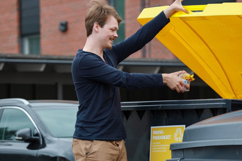 Henkilö nostaa toisella kädellä muovipakkausten keräysastian kantta, toisessa kädessä muovipakkauksia. Taustalla näkyy musta auto ja kerrostalon tiiliseinä.