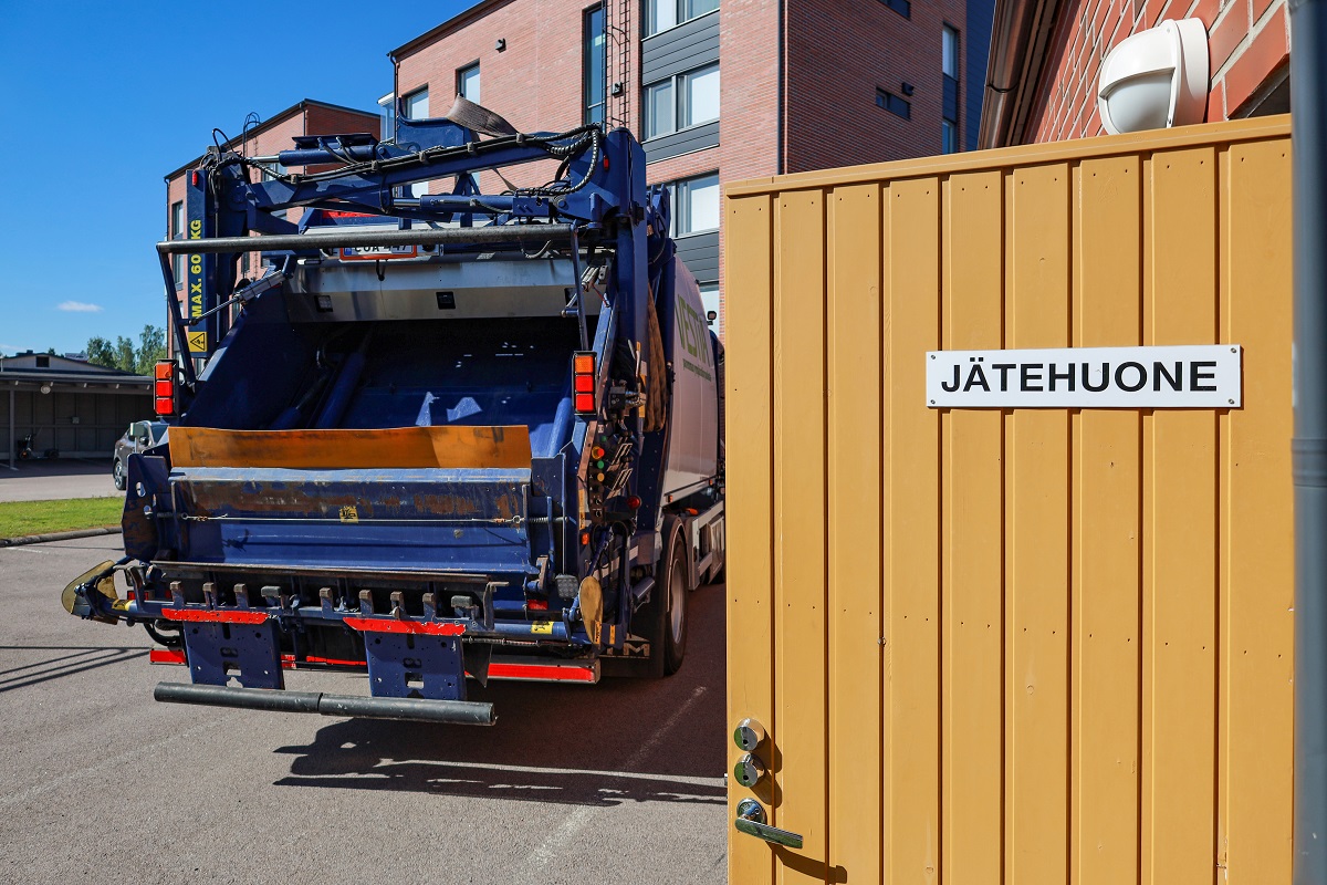 Oikeassa reunassa jätehuoneen avoinna oleva ovi ja vasemmalla jäteauton perä. Taustalla näkyy kerrostalo ja taivasta.