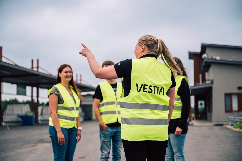 Selkäpuolelta kuvattu Vestian kierrätysneuvoja osoittaa jätelavoille päin ja puhuu kolmelle heijastinliiveihin pukeutuneelle henkilölle Ylivieskan lajittelupihalla. Taustalla näkyy Vestian rakennuksia ja asfaltoitua pihaa.