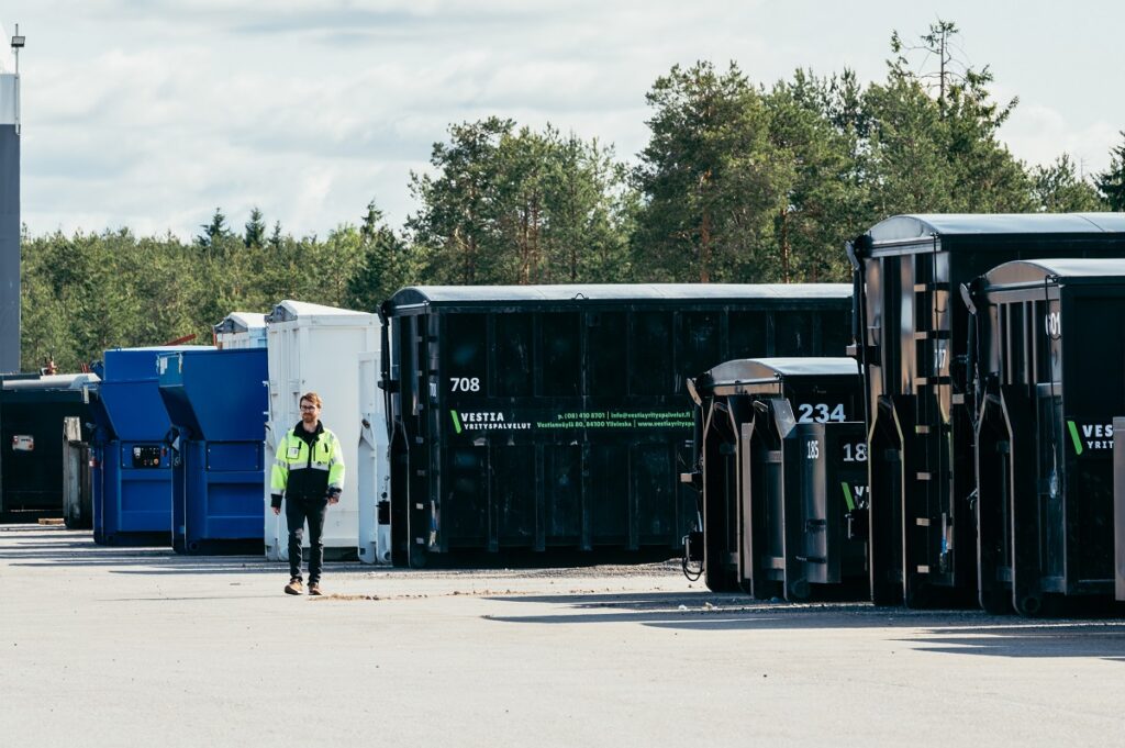 Jätekontteja ja -lavoja rivissä asfaltilla sekä henkilö kävelemässä näiden ohi.