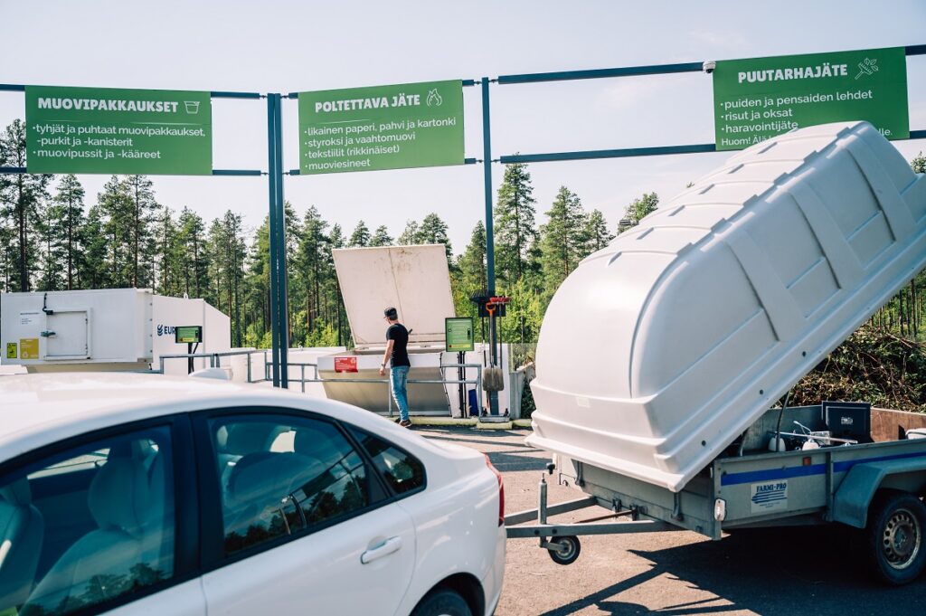 Kuvassa etualalla näkyy valkoisen auton takaosa ja peräkärry kuomu avoinna. Taustalla näkyy henkilö, jätepuristimia ja lajitteluohjekylttejä.