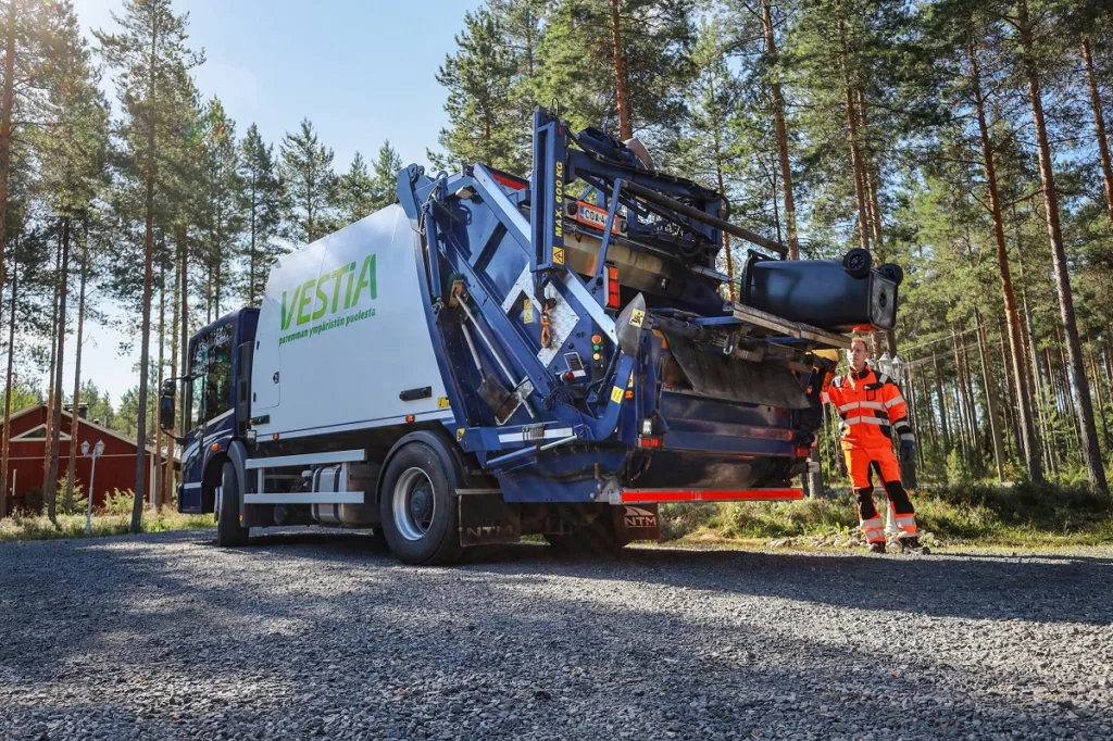 Jäteautonkuljettaja tyhjentämämässä jäteastiaa Vestian jäteautoon. Taustalla metsää ja punainen omakotitalo.